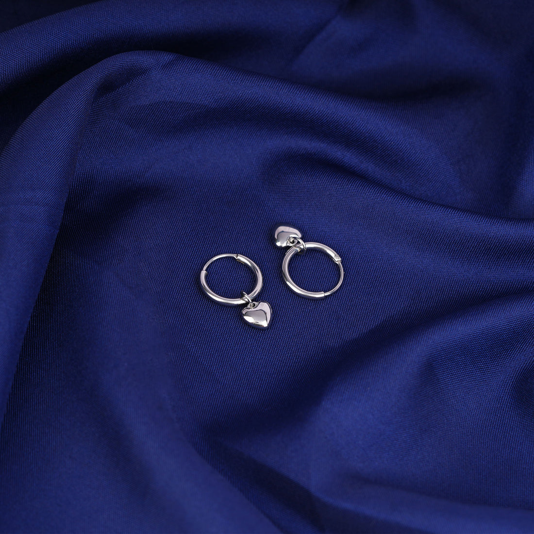 Silver hoop earrings with heart charms on a blue fabric background