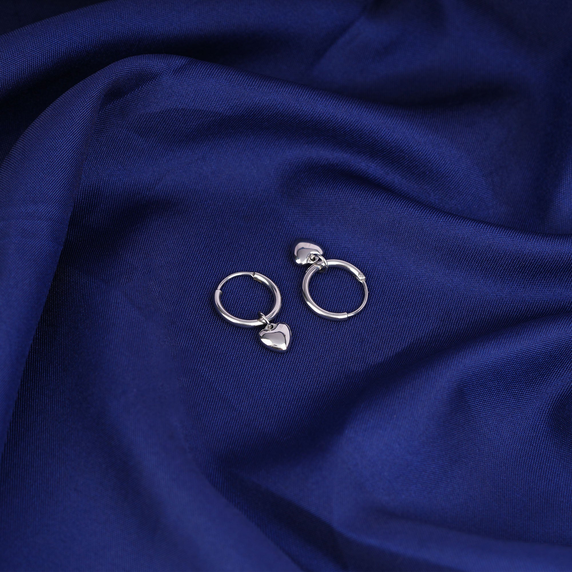 Silver hoop earrings with heart charms on a blue fabric background