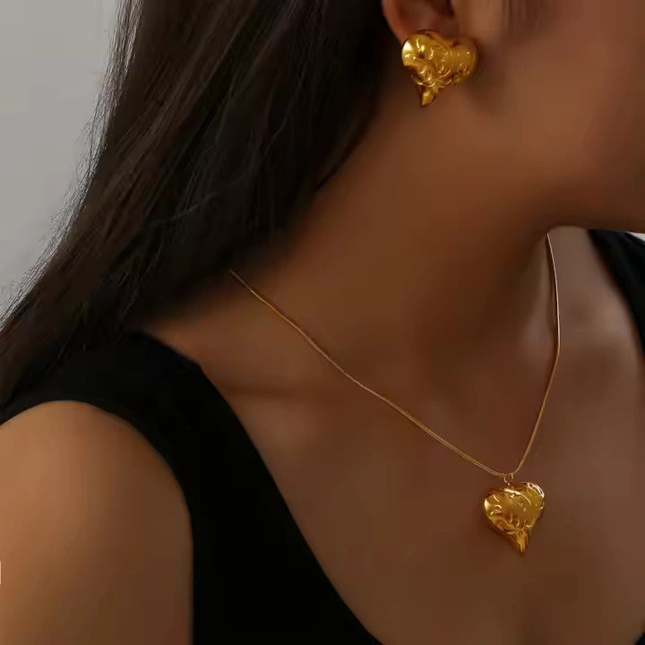 Gold heart-shaped earrings and necklace on a person against a neutral background