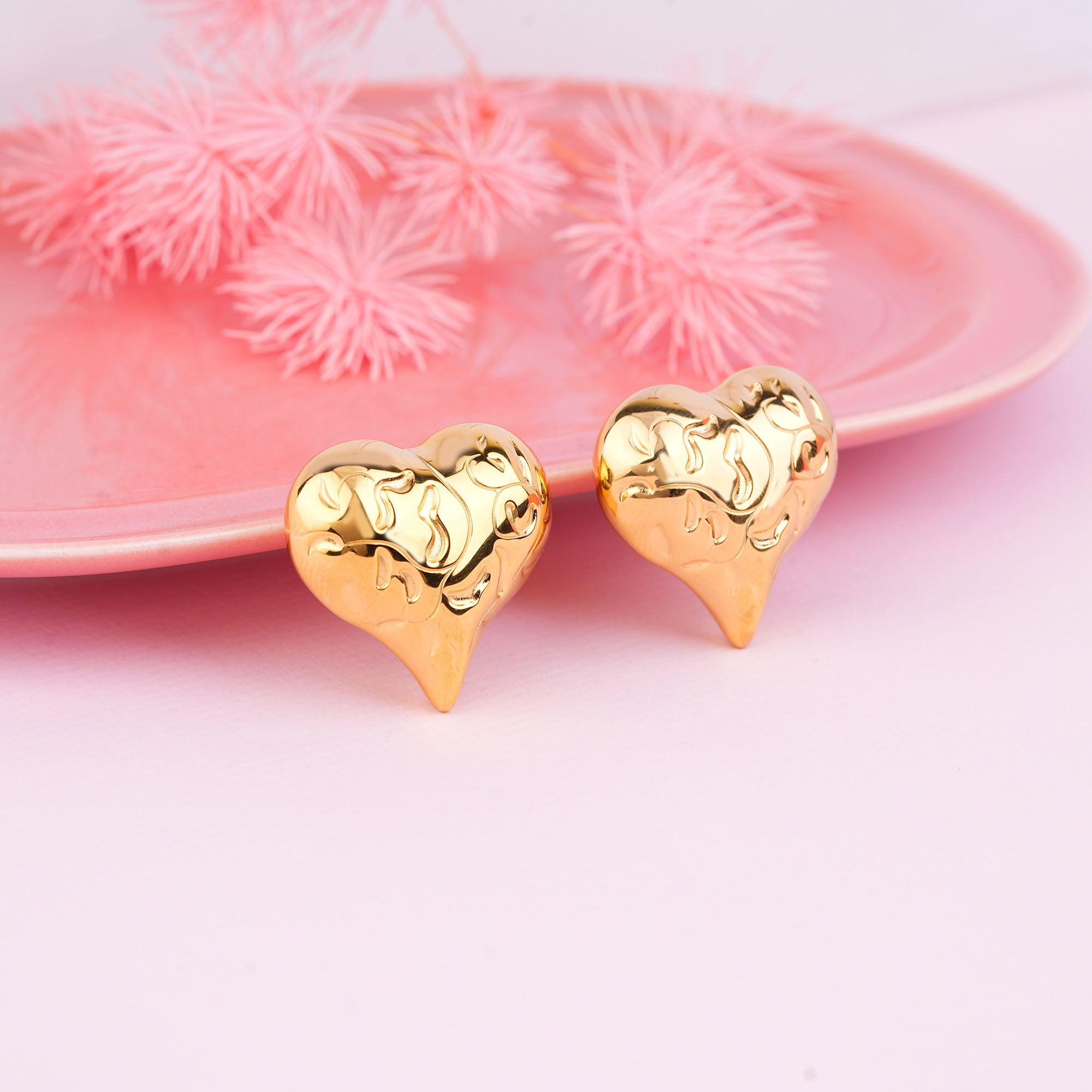 Gold heart-shaped earrings on a pink surface with a decorative plate in the background.